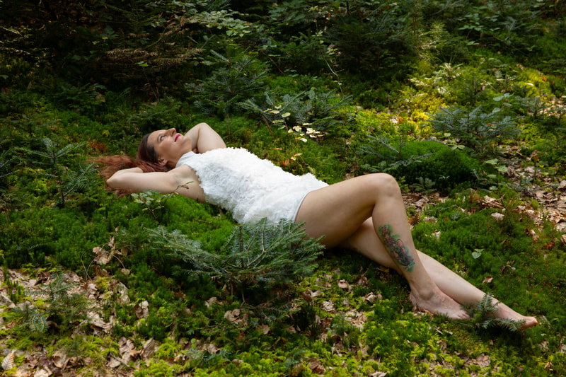 Outdoor- Foto einer Frau in einem weissen Kleid. Sie liegt an einem sonnigen Tag in einem Wald im grünen Moos am Boden